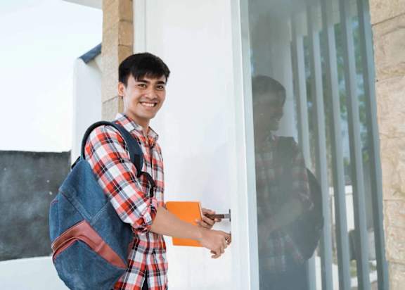 A young man with a backpack and a plaid shirt is holding a book and smiling.