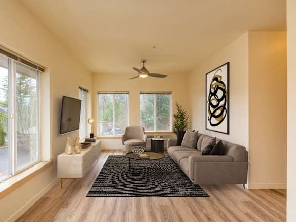 A living room with a grey couch, a television, a ceiling fan, and a large window. at Fairview Village Apartments, Fairview, Oregon