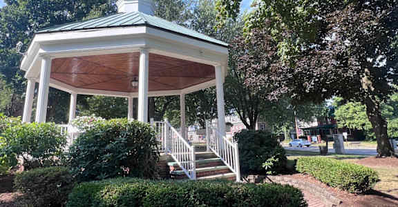 A white gazebo with a green roof is surrounded by trees and bushes.
