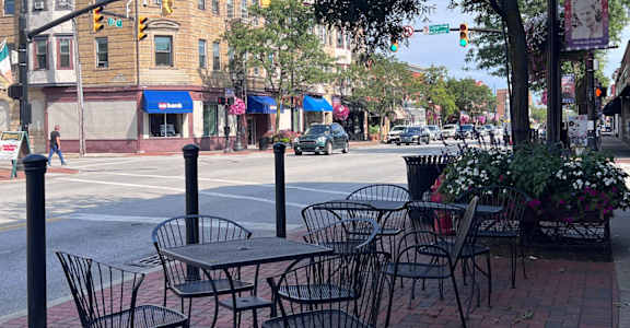 A street view with tables and chairs on the sidewalk.