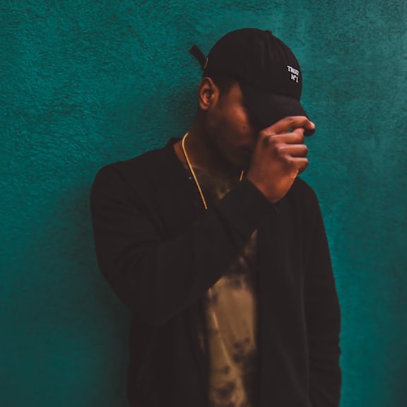 A man in a black jacket and cap is standing against a teal wall.