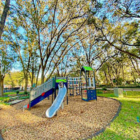 A children's playground on soft wood chips surrounded by grass and large trees near The Flats at Tioga Town Center apartments.