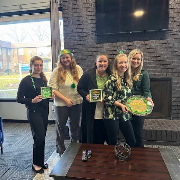 Five women are standing in front of a fireplace holding green themed items.