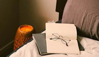 A pair of glasses rests on a book on a bed.