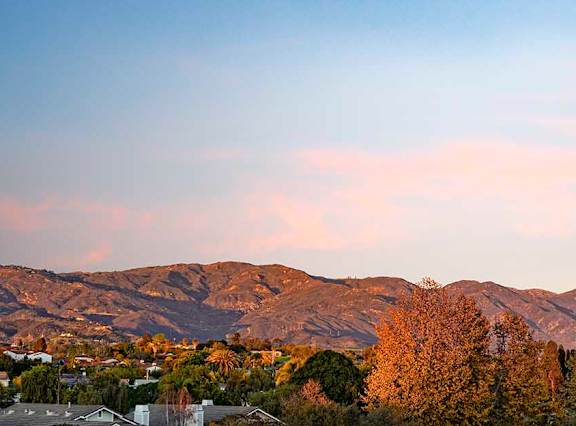 A mountain range with trees in the foreground and houses in the distance under a pink and blue sky.