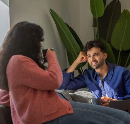 A woman in a pink sweater is taking a photo of a man in a blue shirt who is sitting on a couch with a laptop.