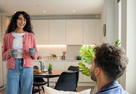 A woman in a pink cardigan and blue jeans stands in a kitchen holding a mug, while a man in a blue shirt sits at a table with a laptop.