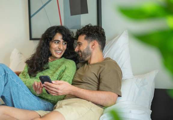 A couple is sitting on a bed, with the man holding a phone and the woman looking at it.