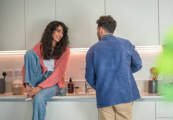 A man and a woman are standing in a kitchen talking to each other.