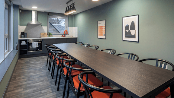 A long wooden table is surrounded by chairs in a room with a kitchen in the background.
