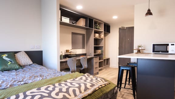 A bedroom with a bed, desk, and kitchenette.