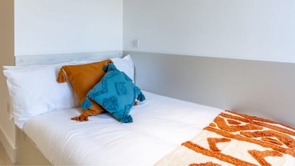 A bed with a white sheet and a blue and orange pillow.