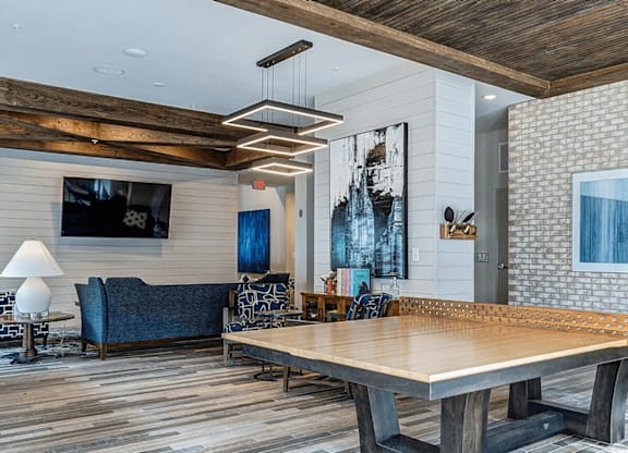 A large wooden table is in the middle of a room with a blue couch and a television mounted on the wall.