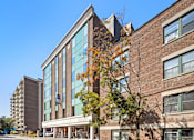 Thumbnail 2 of 4 - A modern building with a glass facade is surrounded by trees with autumn leaves.