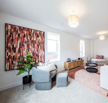 A living room with a grey couch, a grey armchair, a grey ottoman, a wooden coffee table, a pink rug, a large painting on the wall, and a plant.