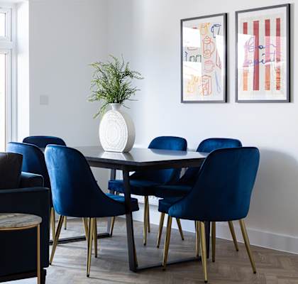 A living room with a dark blue sofa, a round pink chair, and a white vase with a green plant on a black and gold dining table.