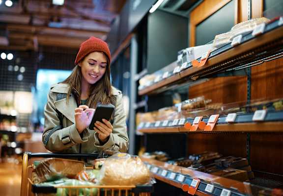 A woman in a beanie is shopping in a grocery store, looking at her phone.