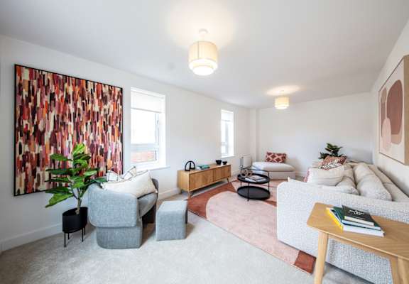 A living room with a grey couch, a grey armchair, a grey ottoman, a wooden coffee table, a pink rug, a large painting on the wall, and a plant.