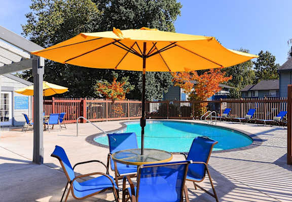 A yellow umbrella shades a table with blue chairs around a pool.