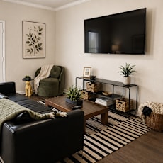 A living room with a black couch, a coffee table, a chair, a basket, and a TV mounted on the wall.