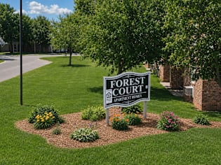 A sign for Forest Court Apartment Homes is in the foreground of a grassy area.