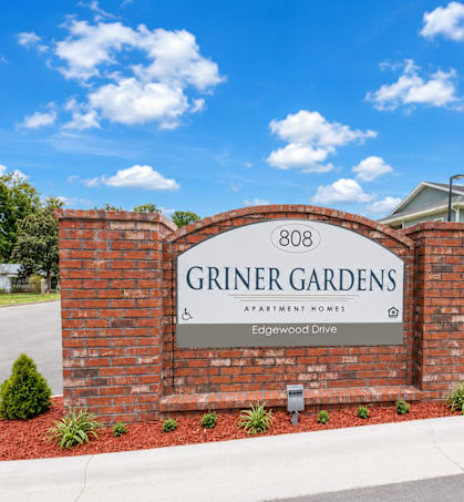 A sign for Griner Gardens Apartment Homes stands in front of a house.