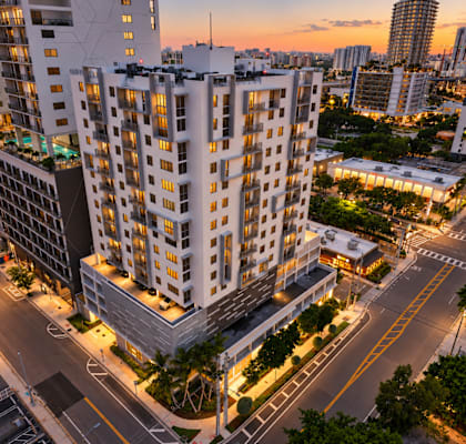 A modern apartment building is lit up at dusk with the sun setting in the background.