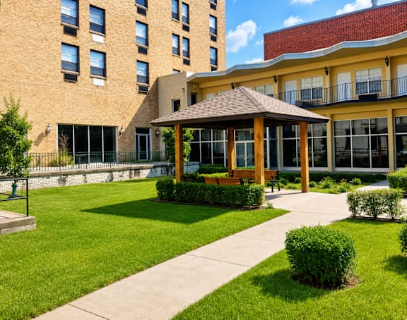 A gazebo is in the middle of a grassy area in front of a tan building.