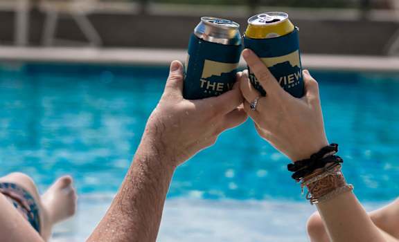 Two people toasting with cans of beer by a pool.