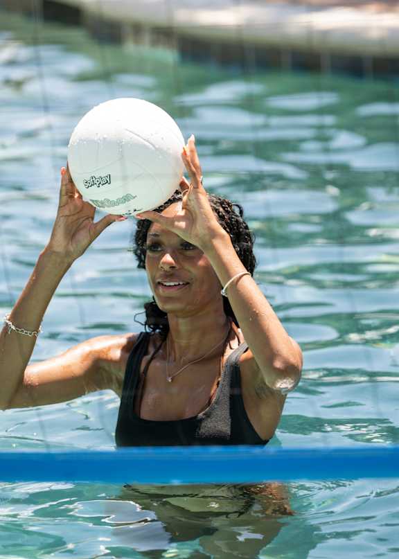 A woman in a black top is holding a white water polo ball up to her face in a pool.