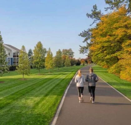 Two people walking down a path in a park with trees and houses in the background.