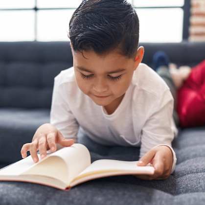 A young boy is reading a book on a grey couch.