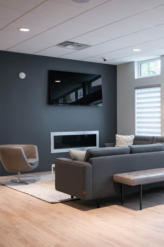 A grey couch with a pillow and a grey chair in front of a grey wall with a tv.