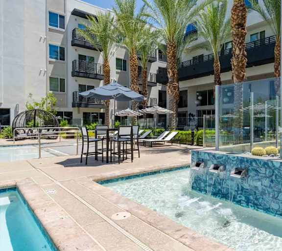 A pool area with a hot tub and lounge chairs in front of a building with palm trees.