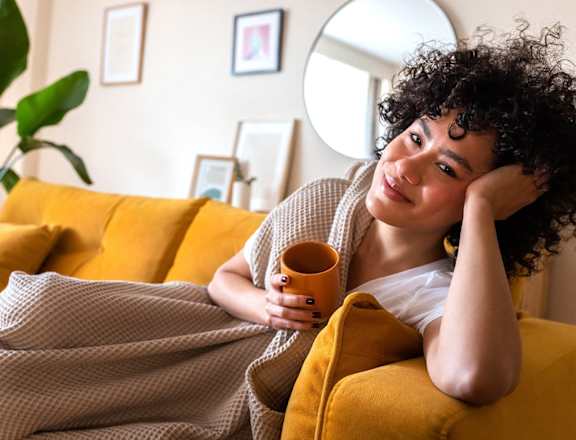a woman laying on a couch under a blanket holding a coffee cup