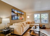 Thumbnail 4 of 34 - Apartments Near Happy Valley For Rent - Sunnyside Village - a Living Room with Wood-Style Flooring, a Large Plush Couch, a Coffee Table, a Small Table with a Lamp, Large Windows with White Blinds, and a Door to the Private Patio