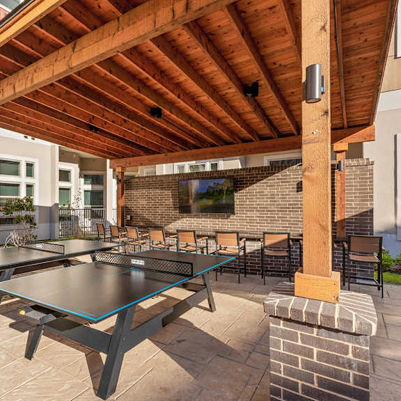 The outdoor lounge area with a covered area, barstools at a long table in front of a TV mounted on a brick wall, and two tables for table tennis at Waterford Market Apartment Homes. 