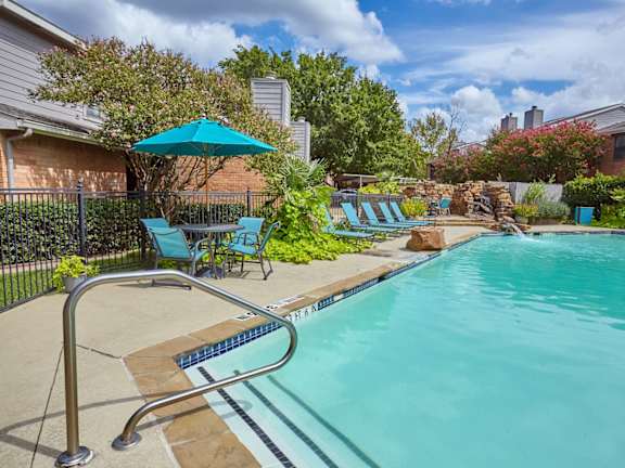 Swimming pool at Fusion Crossroads Apartments with blue lounge chairs, blue umbrellas, and brick buildings in the background.