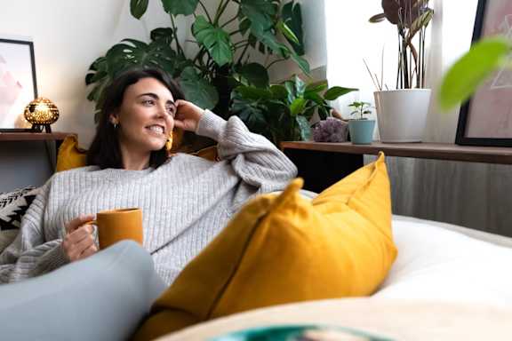 A woman is sitting on a couch in a living room with a bed, a table, and a window.