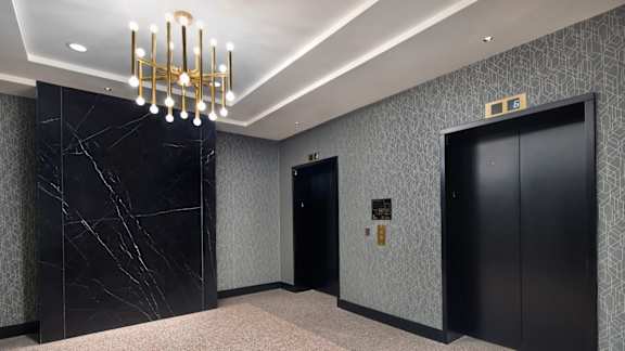 A hallway with a chandelier, marble wall, and two elevators.