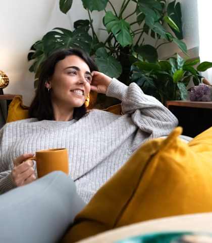 A woman in a grey sweater is sitting on a bed with a yellow pillow, holding a cup, and smiling.