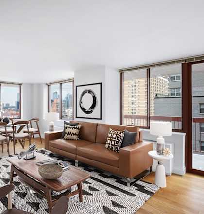 A living room with a brown couch, a coffee table, and a large window overlooking the city at The Ellington Apartments, New York, NY, 10019