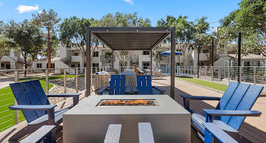 A fire pit is surrounded by blue chairs and benches in a sunny outdoor setting.