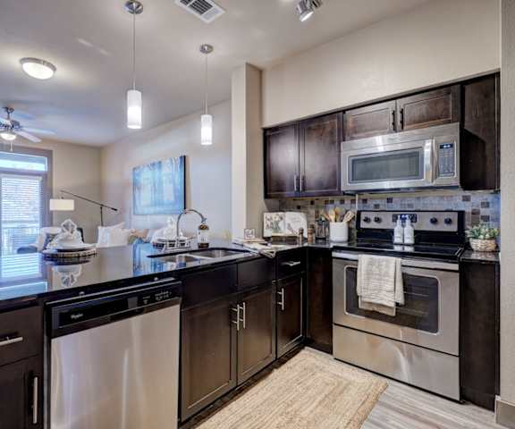 A large kitchen with stainless steel appliances, black bar counter with barstools, kitchen utensils, black countertops with lots of room, and large dark wooden cabinets at the Edge at Midtown Apartment Homes. 
