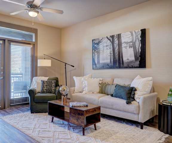 The spacious living room with a white couch, throw pillows, large windows, a ceiling fan, wall art, a green plush chair, coffee table, decorations, large windows, a TV, and glass doors to the balcony at the Edge at Midrown Apartment Homes. 