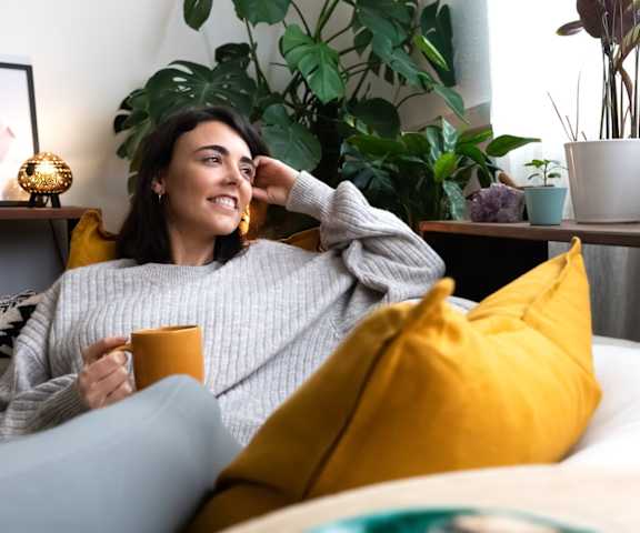 A woman in a grey sweater is sitting on a bed with a yellow pillow, holding a cup, and smiling.
