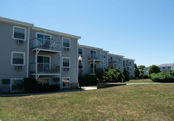 a row of grey apartment buildings with balconies and a grassy area in front of them