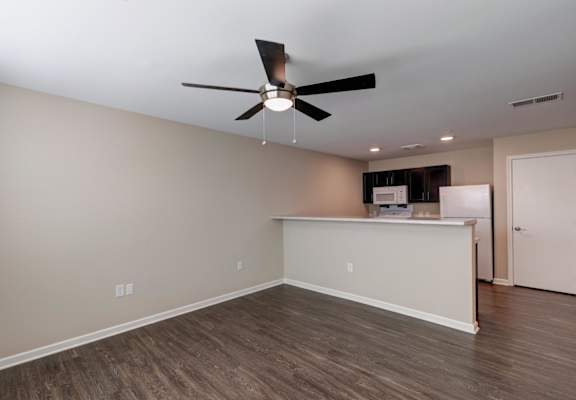 an empty room with a ceiling fan and a kitchen in the background