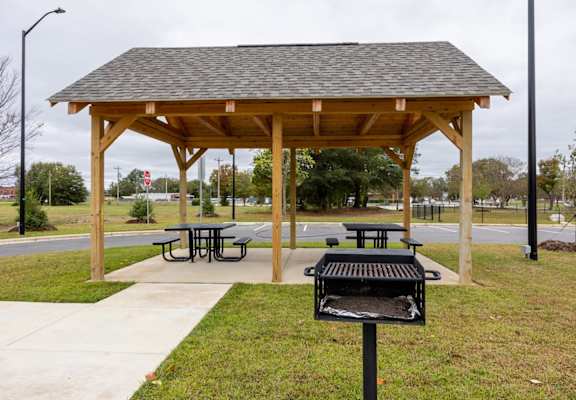 A wooden pavilion with a grill and picnic tables is set up in a grassy area.
