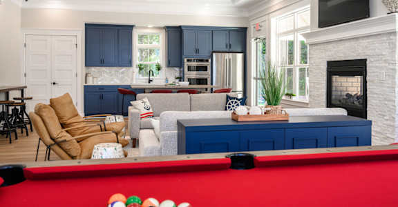 A pool table with a red cloth and white balls is in the foreground of a well-lit room with a fireplace and a television at The Promenade at Easton Apartments, Easton, MD